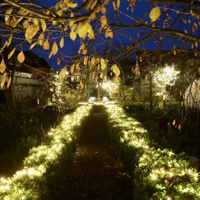 Ein Weg im  Garten des Klosters Wiedenbrück bei Dunkelheit. Der Weg ist umrahmt von kleinen Hecken. Sowohl der Weg als auch der Garten insgesamt werden von zahlreichen Lichtern erleuchtet. © Kloster Wiedenbrück eG.