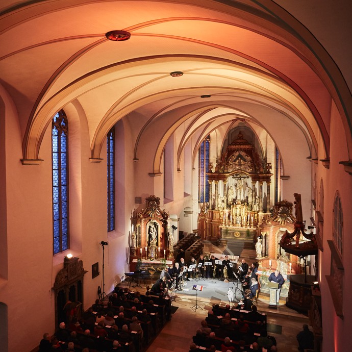Die Kirche des ehemaliges Franziskanerkloster Warendorf von innen. Der Raum ist von warmem Licht ausgestrahlt. Zwischen den beiden Seitenaltären befinden sich Musikanten. Am rechten Seitenaltar befindet sich ein Ambo.© LWL Fotografie Sarah Bömer.